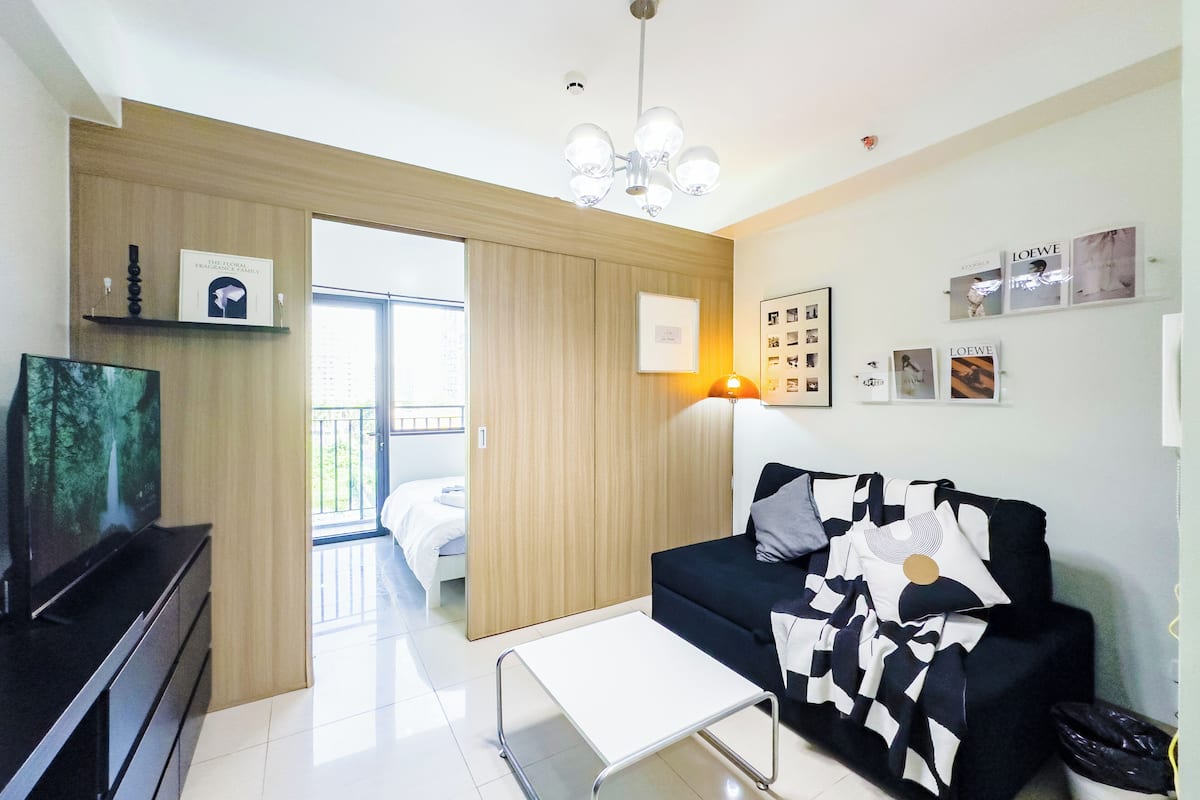 Un salón moderno con un sofá negro, una mesa de centro blanca y un televisor sobre un mueble negro. Hay una puerta ventana que da a un espacio exterior.
