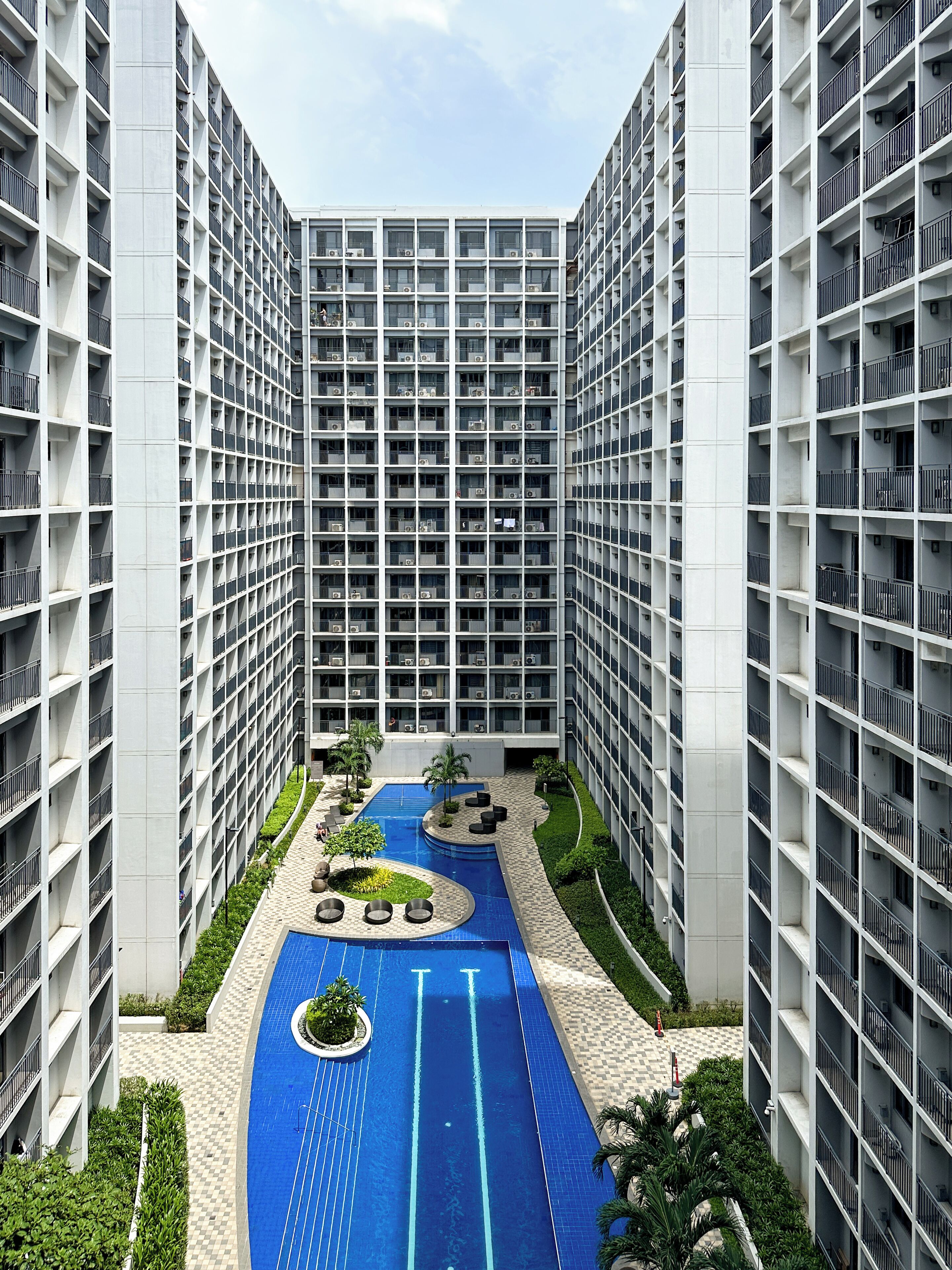 A high-rise building with multiple balconies, a central courtyard with a swimming pool, and surrounding greenery.