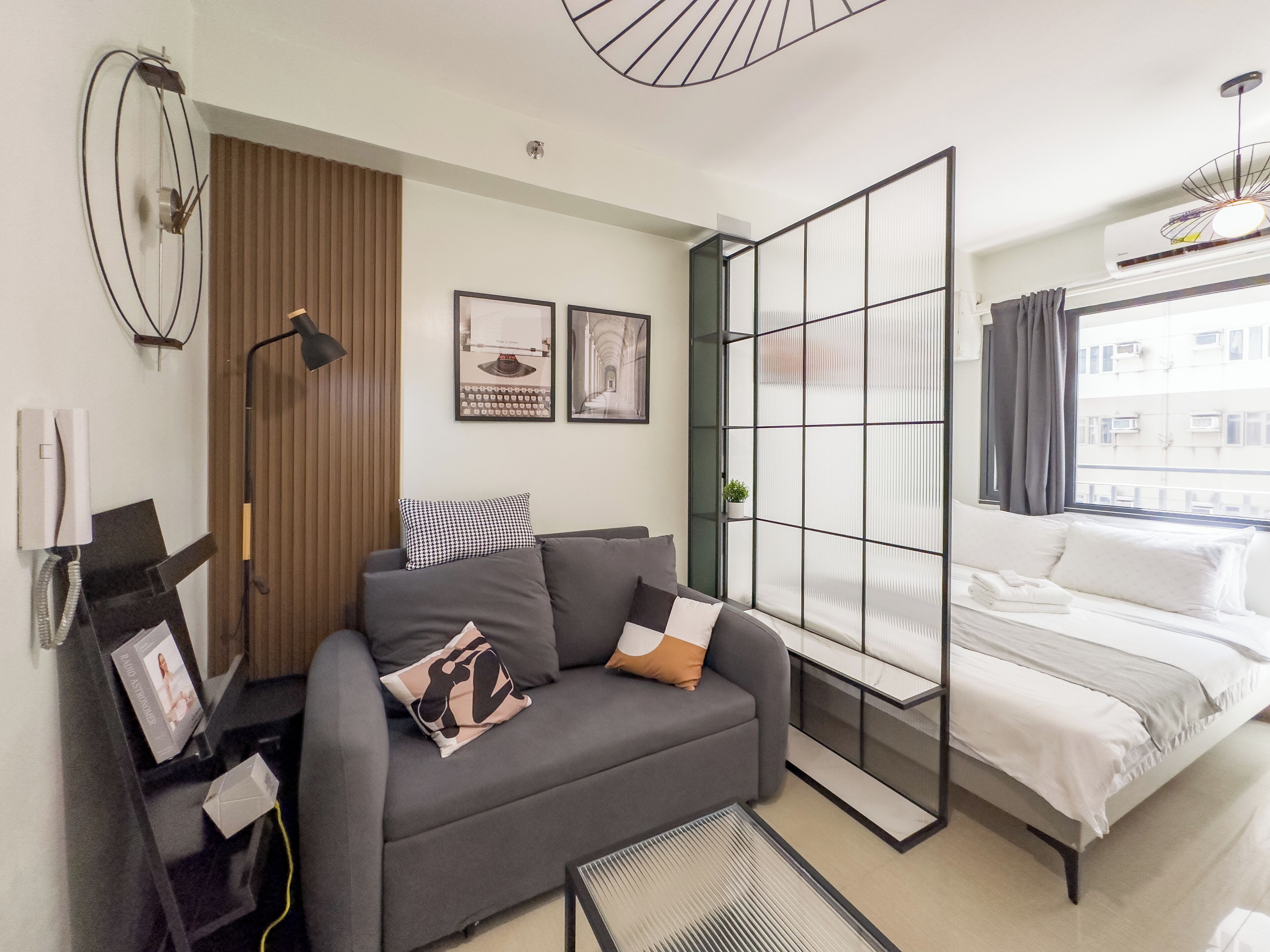 A modern hotel room with a grey sofa, a bed with white bedding, a large window with curtains, a wall-mounted air conditioner, and framed pictures on the wall.