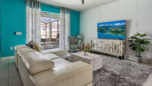 A living room with a teal wall, a ceiling fan, a TV on the wall, a sofa, a coffee table, and a plant. - Magic Beyond The Reef -5BR- Windsor Island- Disney (Davenport)