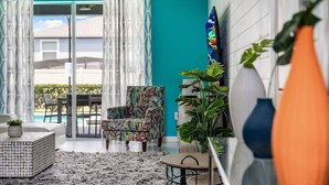 A modern living room with a sofa, a patterned armchair, a coffee table, and a small side table. There is a large window with curtains, a view of the outdoors, and a potted plant. - Magic Beyond The Reef -5BR- Windsor Island- Disney (Davenport)