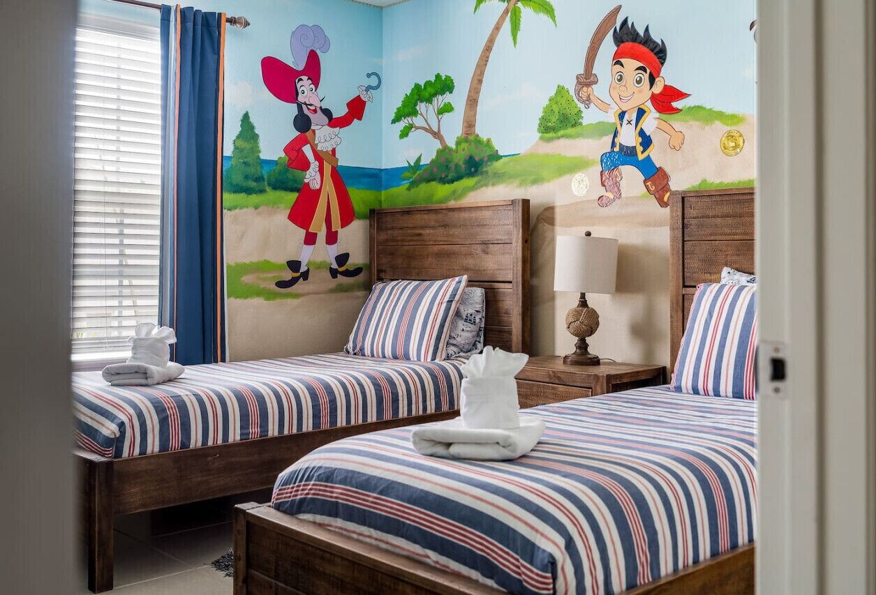 A hotel room with two beds, striped bedding, a mural with cartoon characters, a window with curtains, and a nightstand with a lamp.