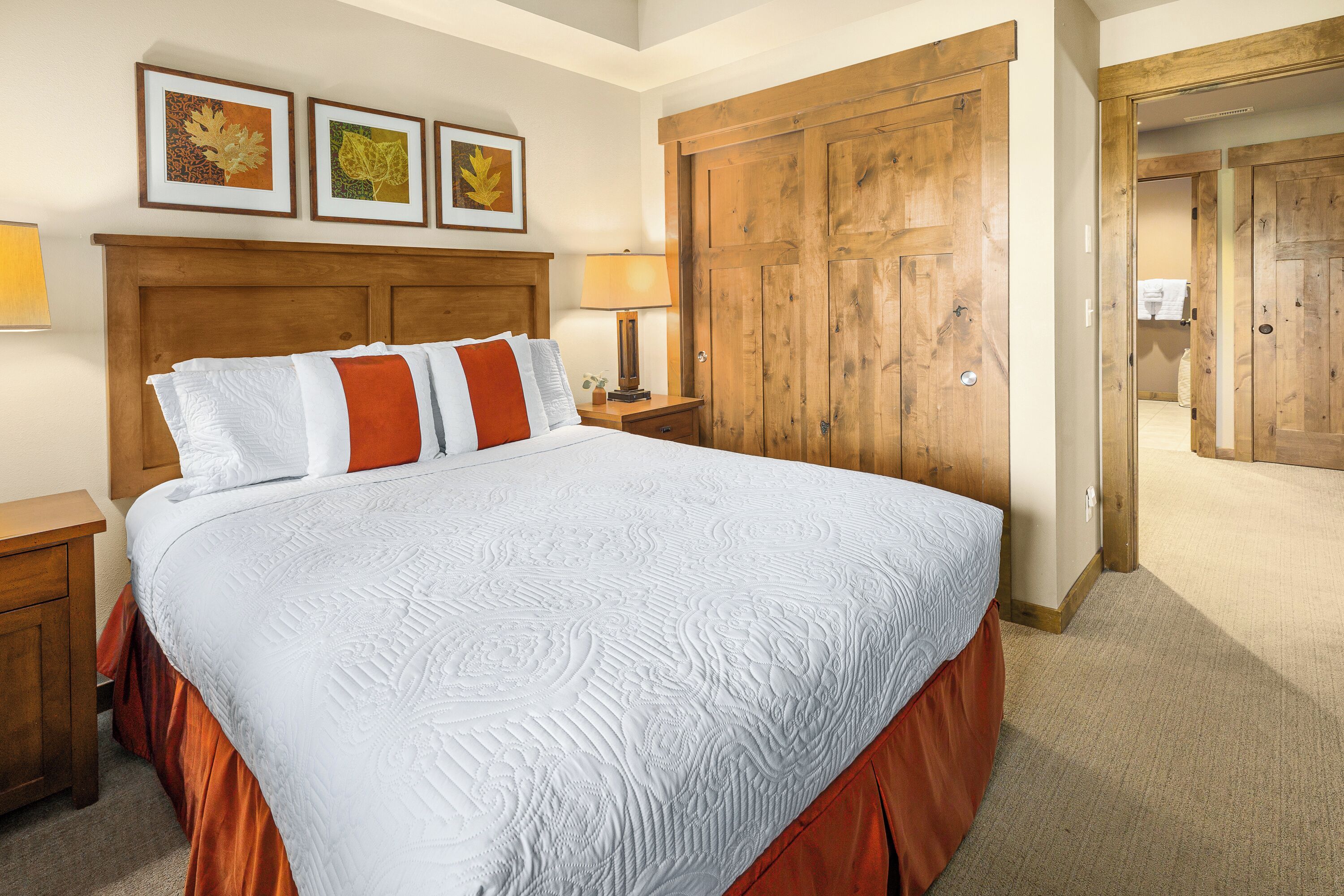 A large bed with a white and orange bedspread, wooden headboard, and two bedside tables with lamps. There are three framed pictures on the wall above the bed.