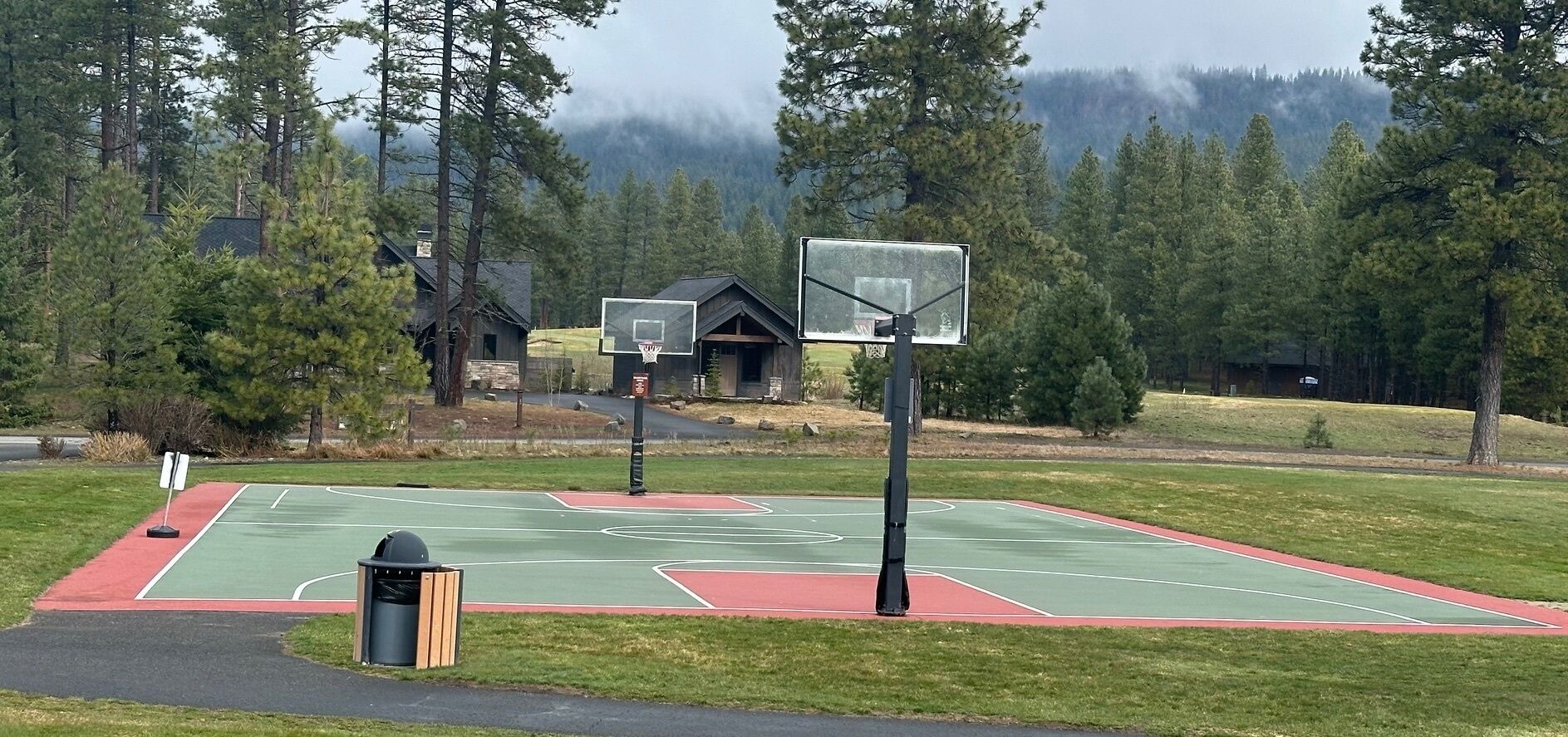 An outdoor basketball court with two hoops, surrounded by trees and a building in the background.