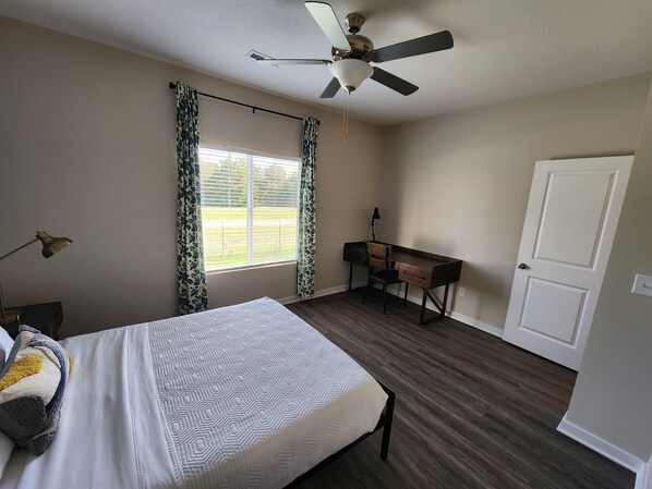 A bedroom with a bed, a desk, a chair, a ceiling fan, and a window with curtains.