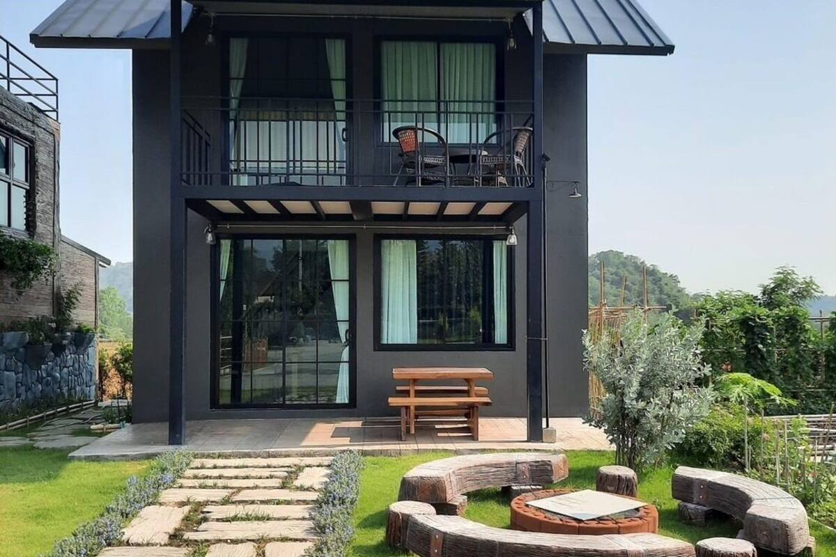A modern two-story house with a balcony, a stone pathway leading to a wooden seating area, and a well-maintained lawn.