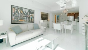 A modern living room with a white sofa, a dining area with white chairs, a kitchen with stainless steel appliances, and a large cityscape painting on the wall.