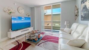 Un saló modern amb un televisió de pantalla plana gran, un sofà blanc, una taula de centre i un balcó amb vista al mar.