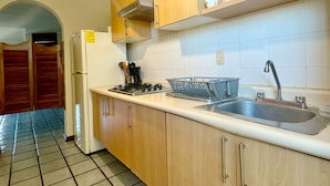 A kitchen with wooden cabinets, a stainless steel sink, and a gas stove. There are jars on a shelf above the sink and a hanging light fixture. - Cozy Apartment Near the Beach (Mazatlán)