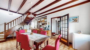 A dining area with a wooden table and chairs, a kitchen in the background, and a staircase leading upstairs.
