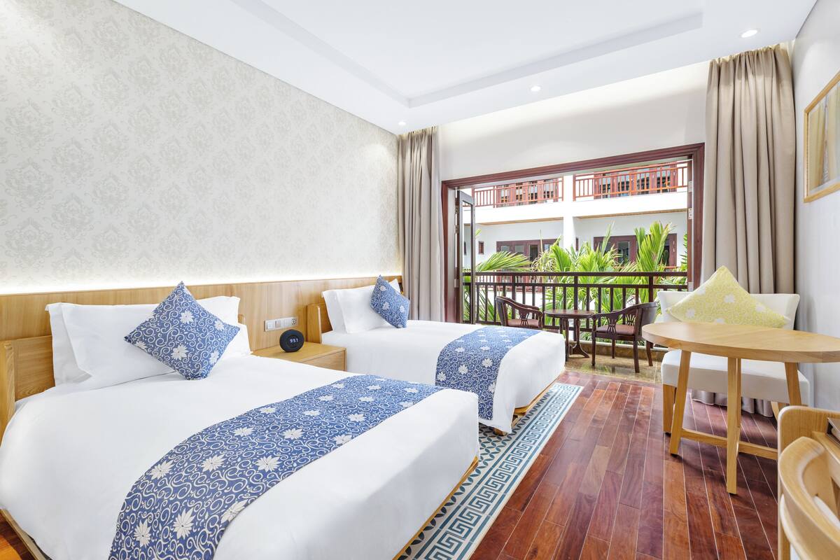A hotel room with two beds, a balcony with a view of greenery, a dining area with a table and chairs, and a patterned rug.