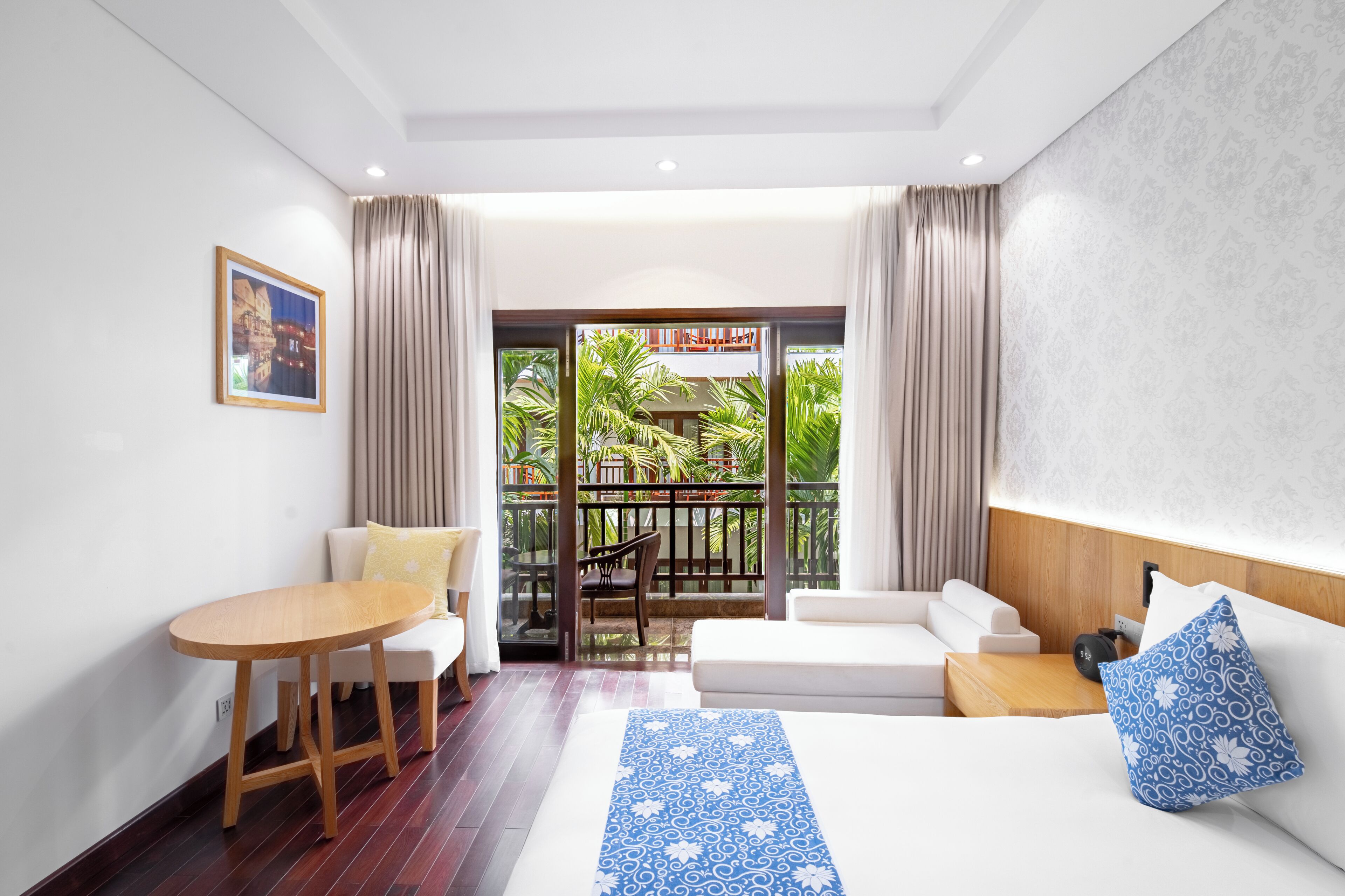 Une chambre d’hôtel avec un lit, une petite table, un canapé, un balcon donnant sur un espace verdoyant et un tableau encadré au mur.