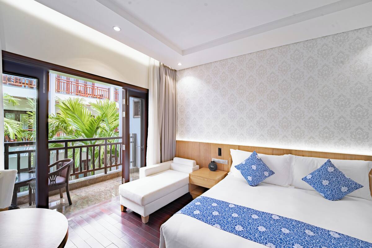 A hotel room with a balcony, a bed with patterned bedding, a wooden nightstand, and a dining area with a table and chairs.