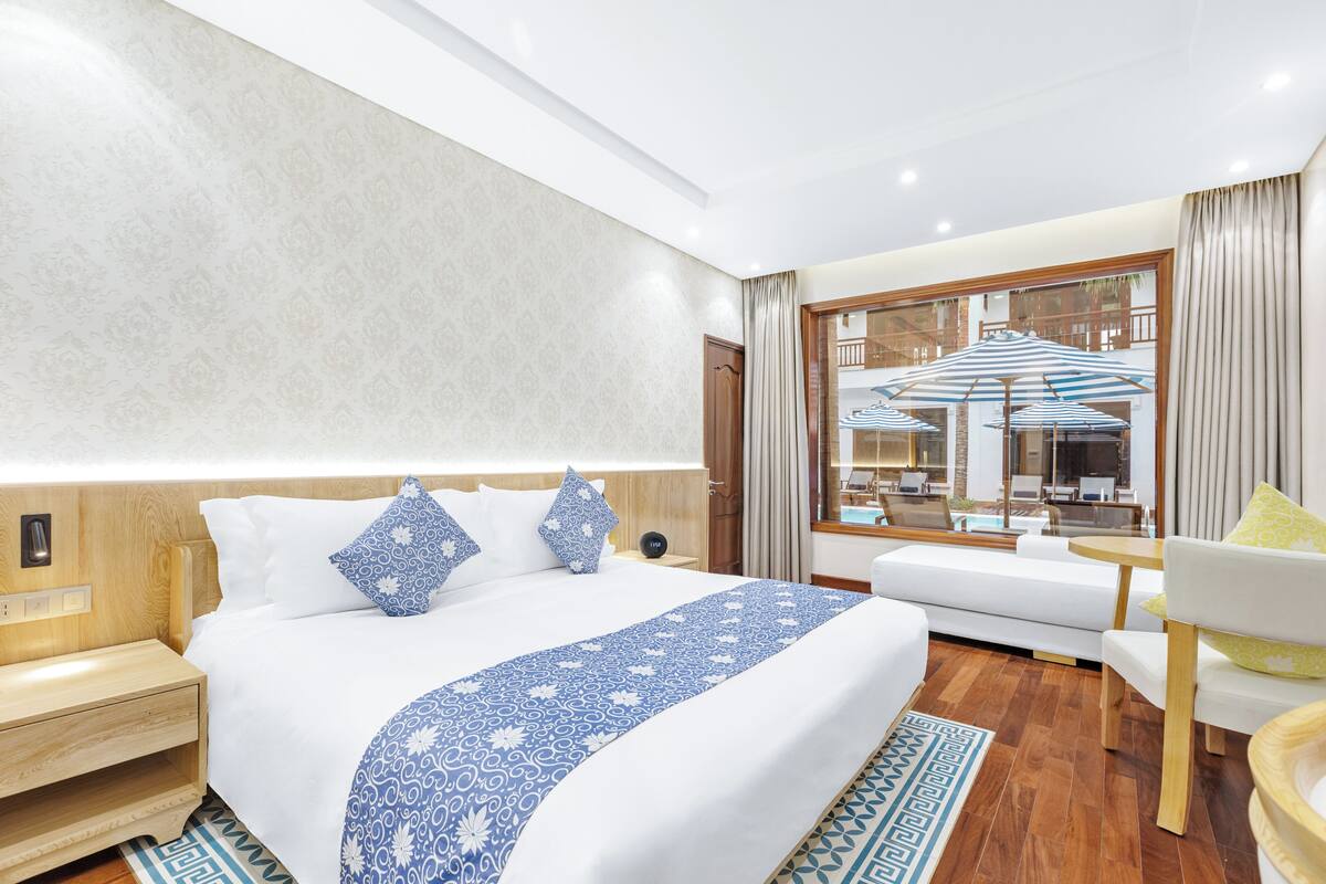 A modern hotel room with a large bed, a sitting area with a sofa and armchairs, a wooden floor, and a view of the pool area through a large window.
