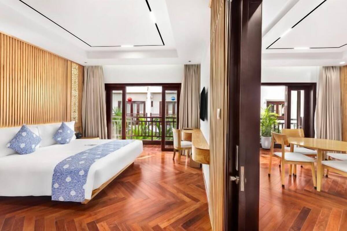 A hotel room with a bed, wooden flooring, a dining area with a table and chairs, and a view of the outdoors.