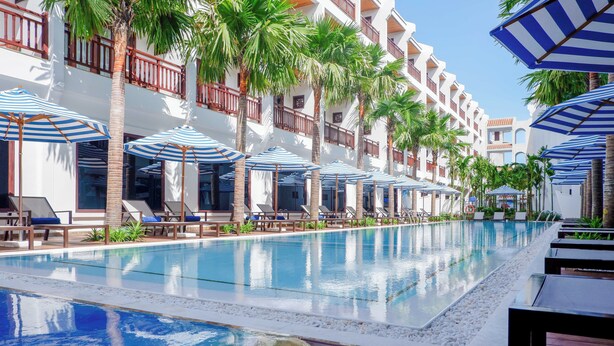 A hotel pool area with multiple lounge chairs and umbrellas, surrounded by palm trees and a multi-story building with balconies. - Wafaifo Resort Hoi An (Da Nang)
