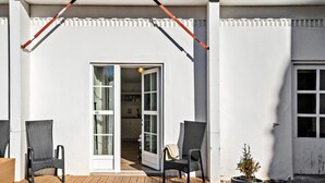 En overdækket terrasse med to fletstole, en potteplante og en skydedør, der fører til et rum indendørs.