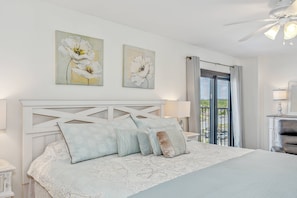 A bedroom with a large bed, a white headboard, two wall-mounted flower paintings, a balcony with a view, and a ceiling fan.