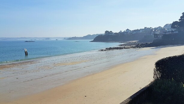Una playa con arena, aguas tranquilas y edificios al fondo.