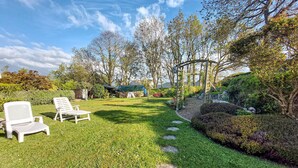 Un jardín bien cuidado con césped, una pérgola de madera y dos sillones blancos.