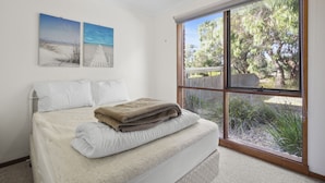 A bedroom with a bed, a window with a view of trees and a fence, and two wall-mounted paintings. - Holiday on Minifie (Anglesea)