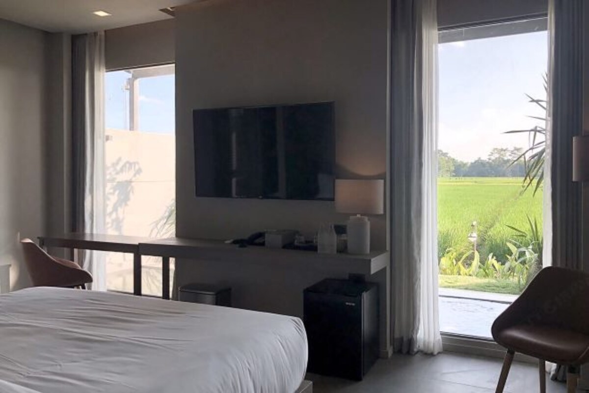 Hotelkamer met een bed, bureau, stoel, televisie en uitzicht op een groen veld door grote ramen.