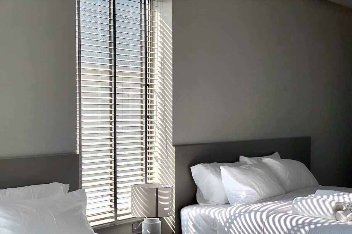 A hotel room with two beds, a large window with blinds, and a bedside table with a lamp.
