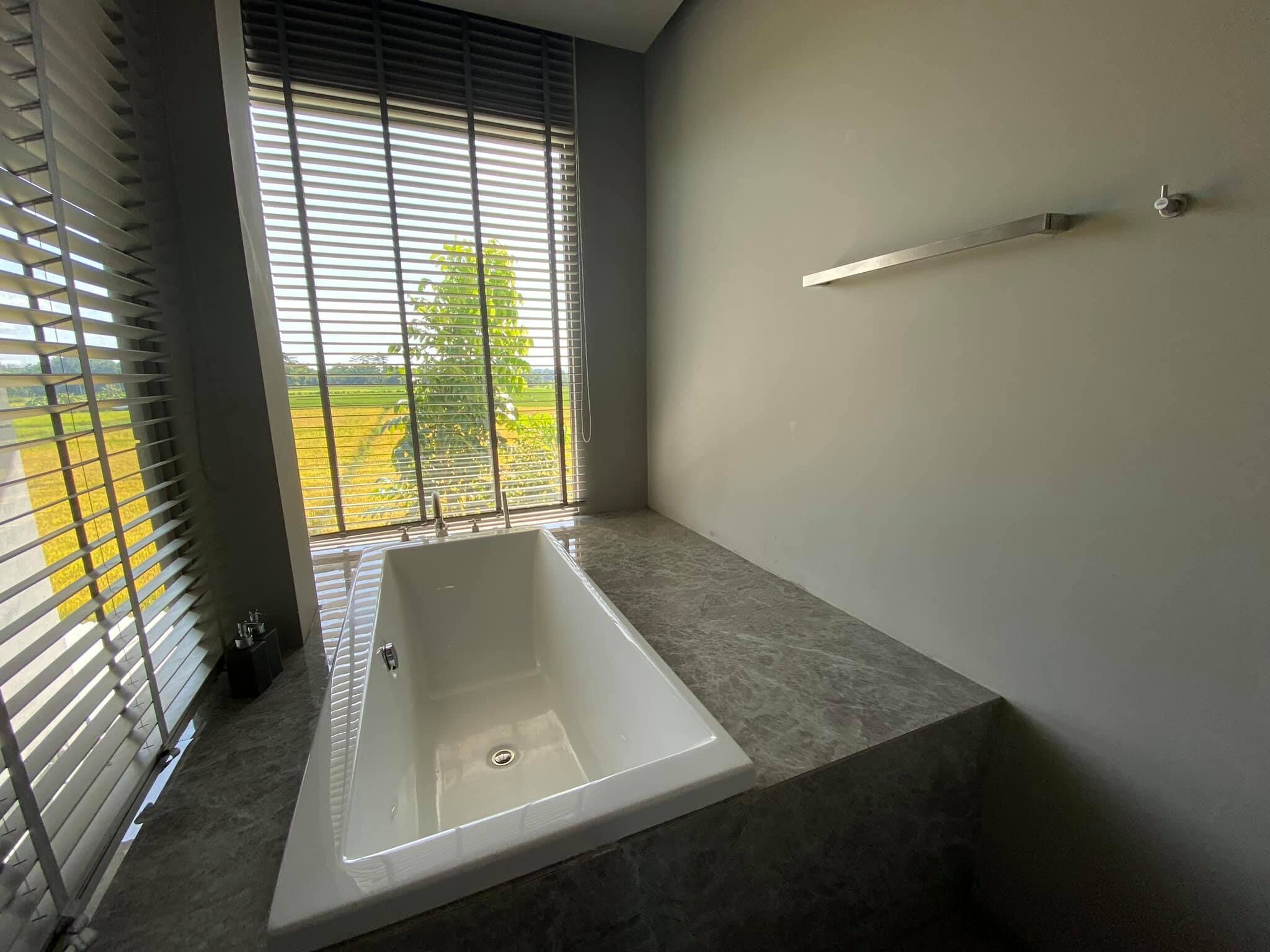 Een moderne badkamer met een badkuip, grote ramen met zonwering en uitzicht op een groen landschap.