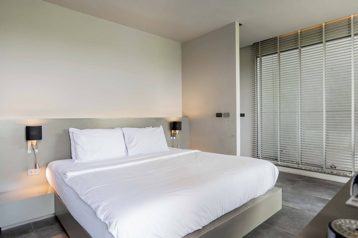 A modern bedroom with a large bed, two bedside tables, and a window with blinds.