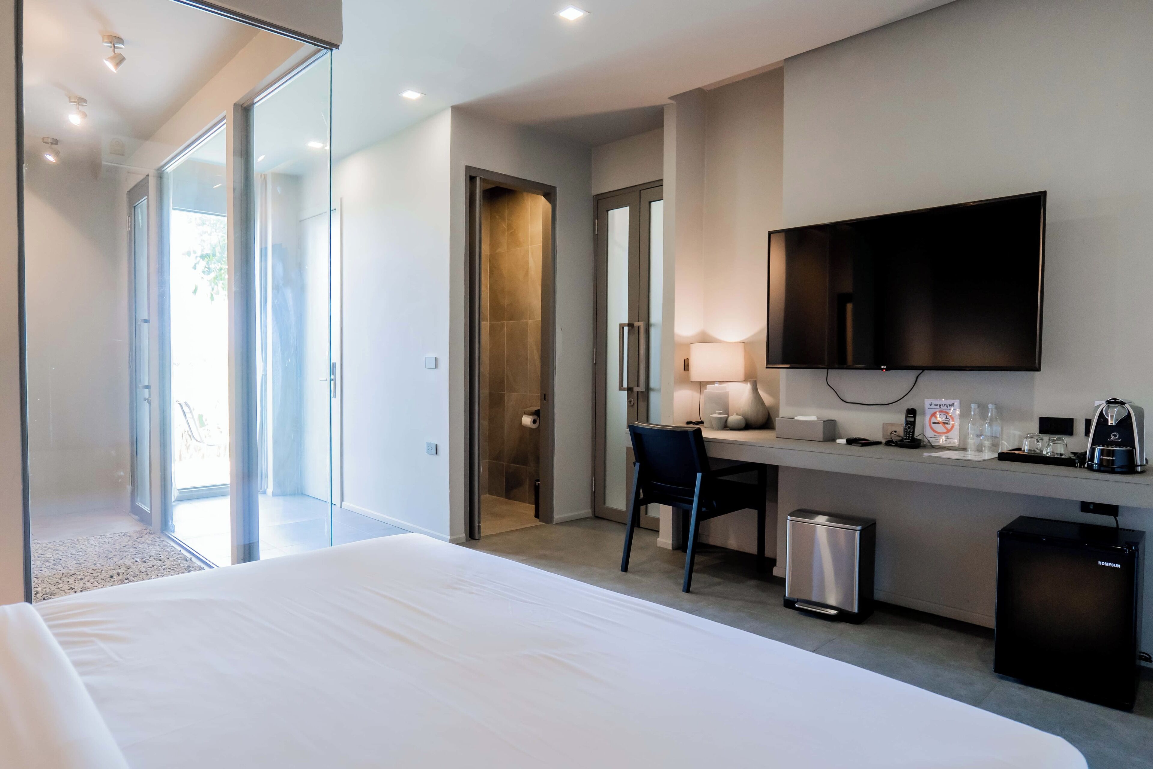 Habitación de hotel con cama, escritorio con silla, televisor, refrigerador pequeño, cesto de basura y puerta corrediza de vidrio que da a un balcón.