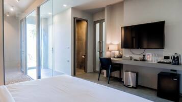 Habitación de hotel con cama, escritorio con silla, televisor, refrigerador pequeño, cesto de basura y puerta corrediza de vidrio que da a un balcón.