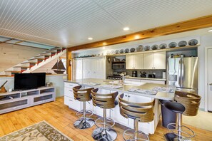 A modern kitchen with white cabinets, a stainless steel refrigerator, a microwave, and a wooden staircase. - Historic Friendship Boat House Near Back River! (Friendship)