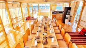 Une longue table de salle à manger en bois, dressée pour un repas, entourée de chaises, avec vue sur l’eau.