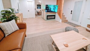 Ein modernes Wohnzimmer mit einer braunen Ledersofa, einem Holztisch und einem an einer Holzwand montierten Fernseher. Im Hintergrund ist eine Essecke mit Tisch und Stühlen zu sehen, und eine Treppe führt zu einer weiteren Ebene.