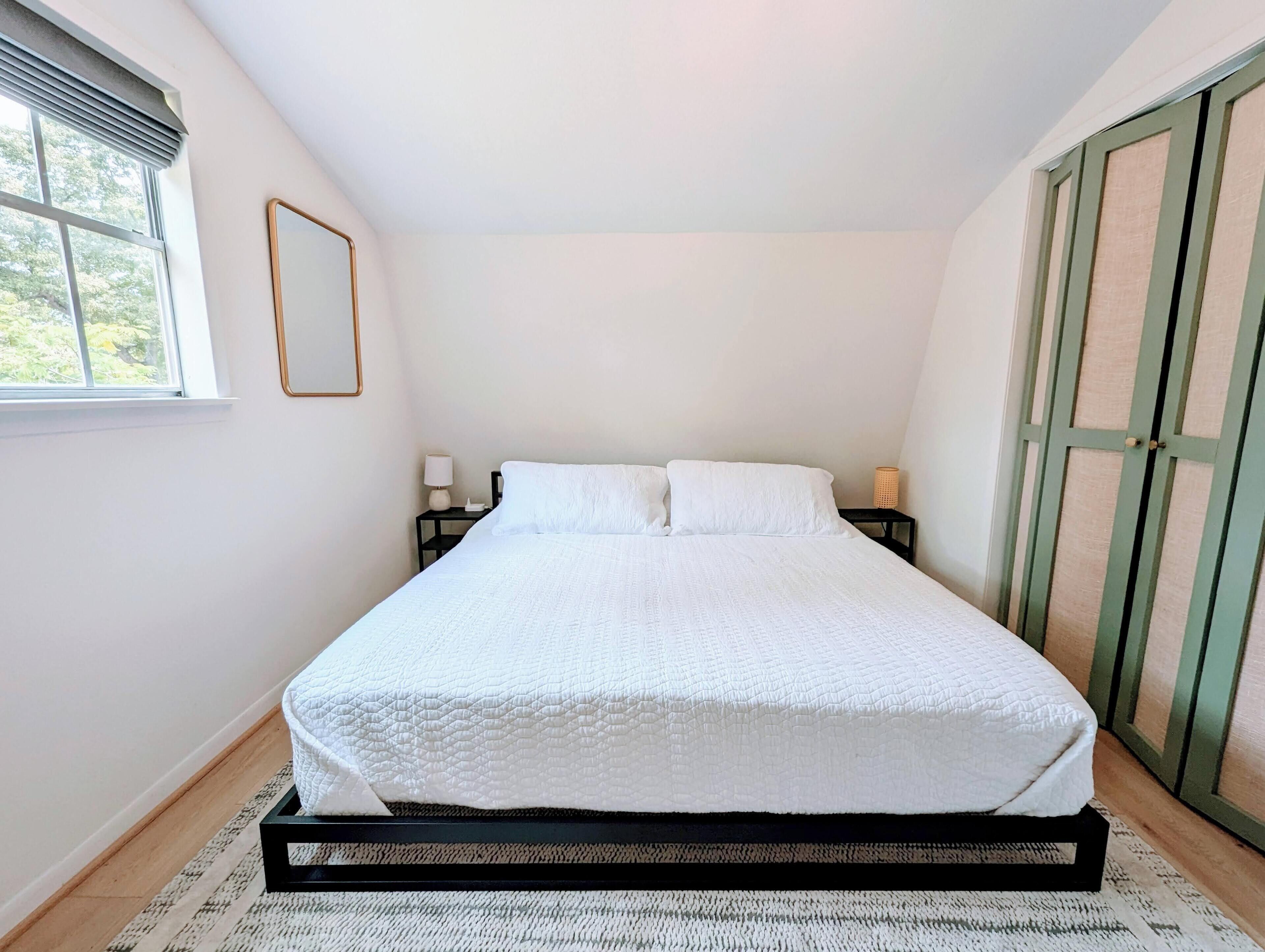 A neatly made bed in a room with a window, a bedside table with a lamp, and a green door.