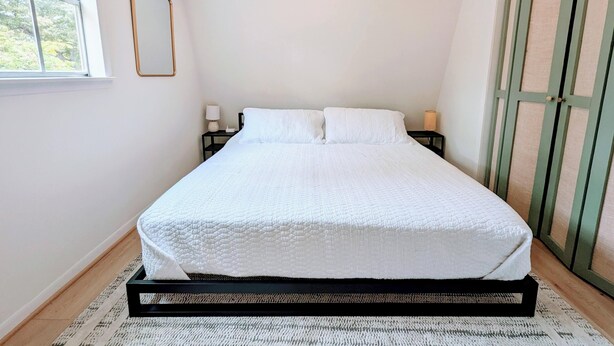 A neatly made bed in a room with a window, a bedside table with a lamp, and a green door.