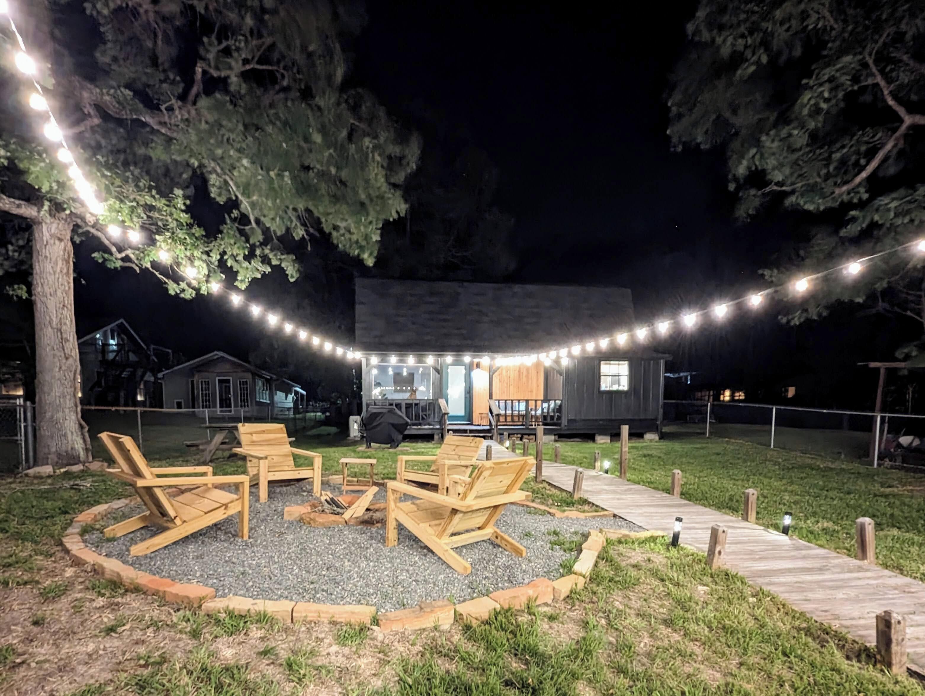 Zona salotto esterna rustica con sedie di legno e un braciere, circondata da un pontile in legno e luci decorative.