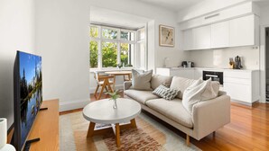 A modern living room with a sofa, coffee table, and TV. There is a kitchen area with cabinets and a sink. Large windows provide natural light and a view of greenery outside. - Inner-West Studio in a Perfect Location (Lewisham)