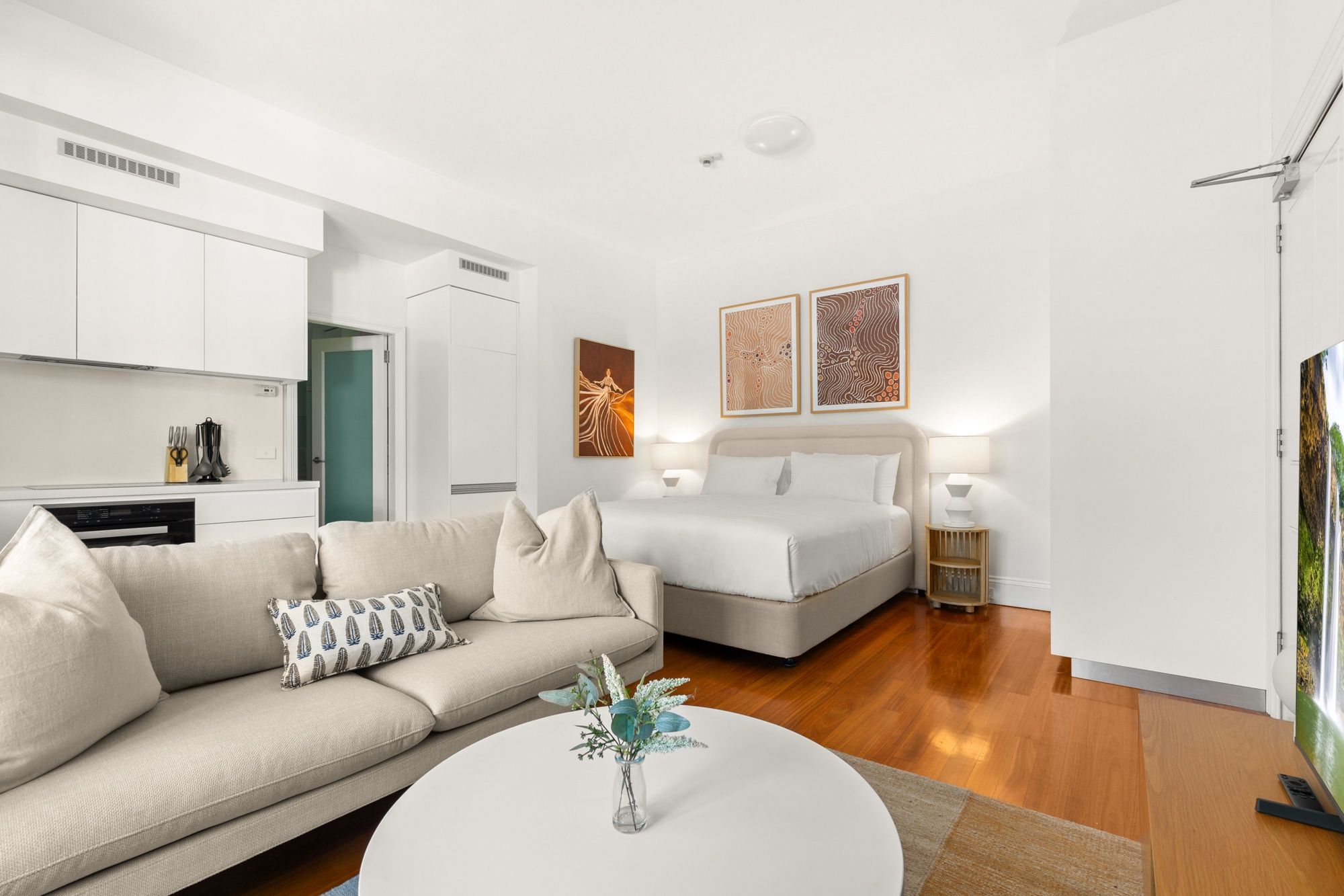 A modern hotel room with a sofa, a bed, a small table with a vase, and artwork on the walls.
