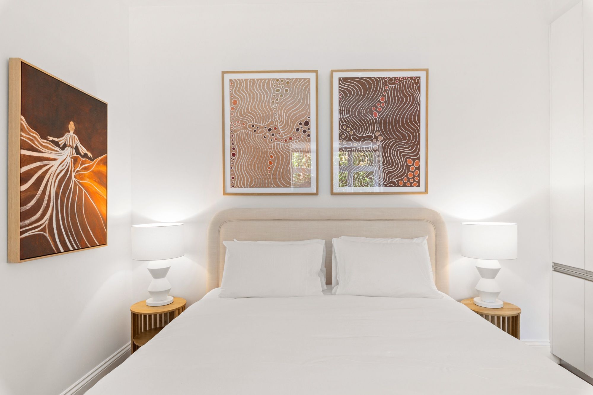 Ein Hotelzimmer mit einem großen Bett, zwei Nachttischen mit Lampen und zwei abstrakten Bildern an der Wand.