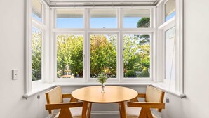 A dining area with a round wooden table and four wooden chairs, a white window with multiple panes, and a view of trees and a fountain outside. - Inner-West Studio in a Perfect Location (Lewisham)