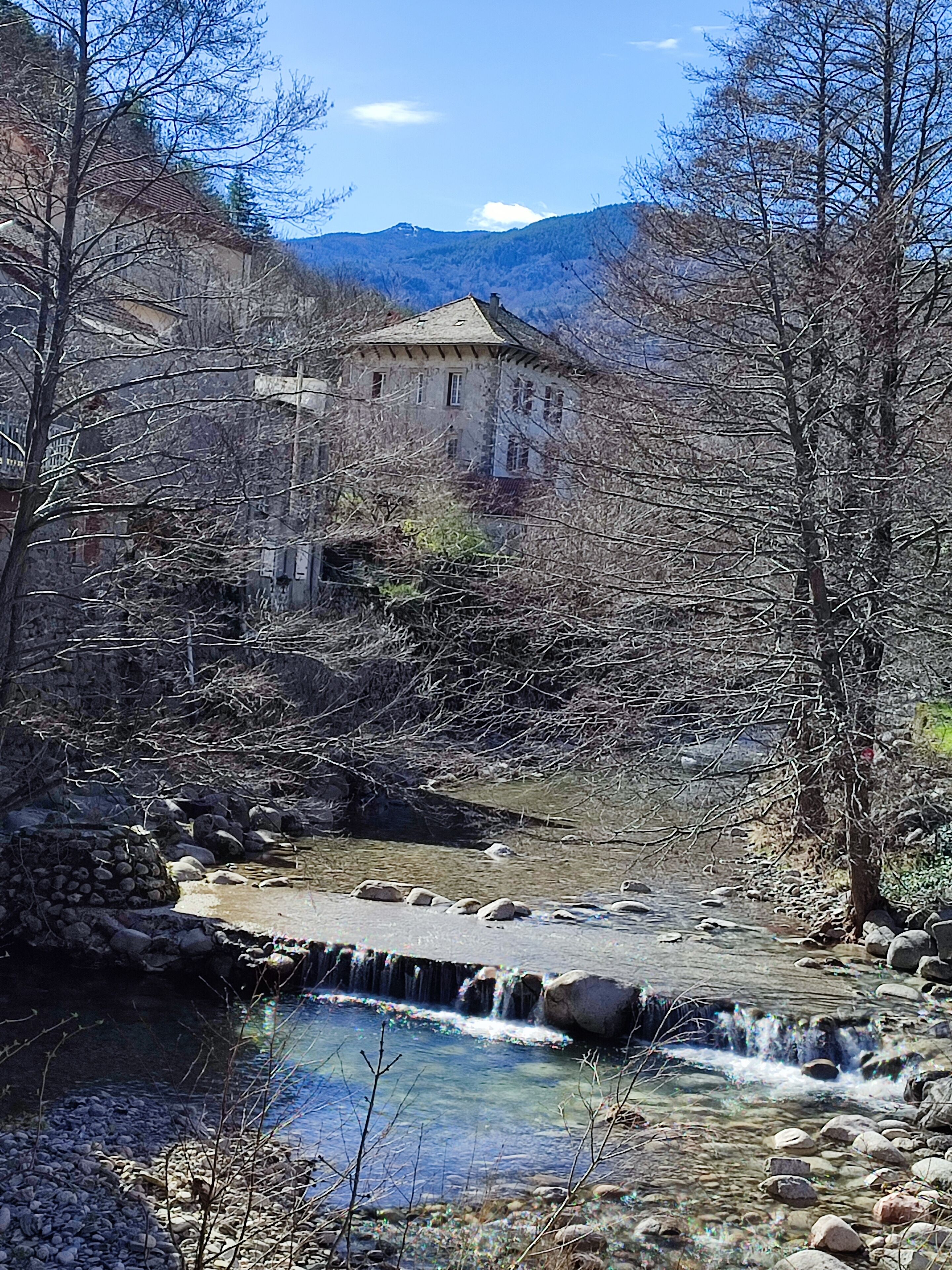 Ein Steinhaus mit einem kleinen Wasserfall davor, umgeben von kahlen Bäumen und einem klaren Himmel.