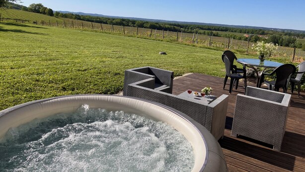 A hot tub on a wooden deck with outdoor furniture and a table, overlooking a vineyard and a grassy field. - Secluded Périgourdine, nestled in the vineyards with a spa and a view (Pomport)