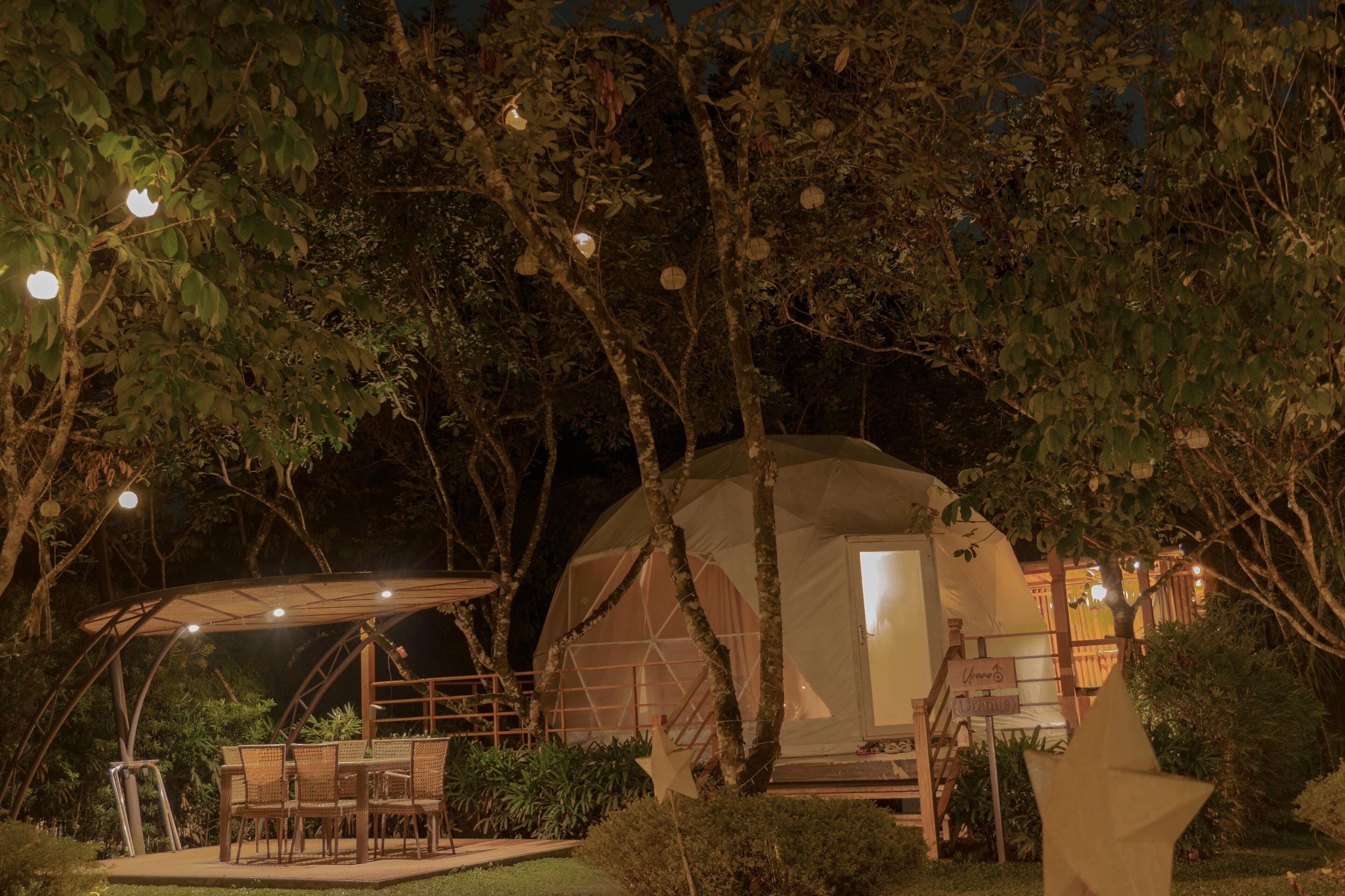 A dome-shaped tent with a staircase leading to the entrance, surrounded by trees and a patio area with chairs and a table.