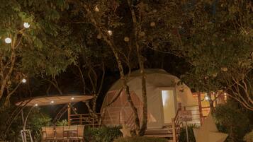 A dome-shaped tent with a staircase leading to the entrance, surrounded by trees and a patio area with chairs and a table.