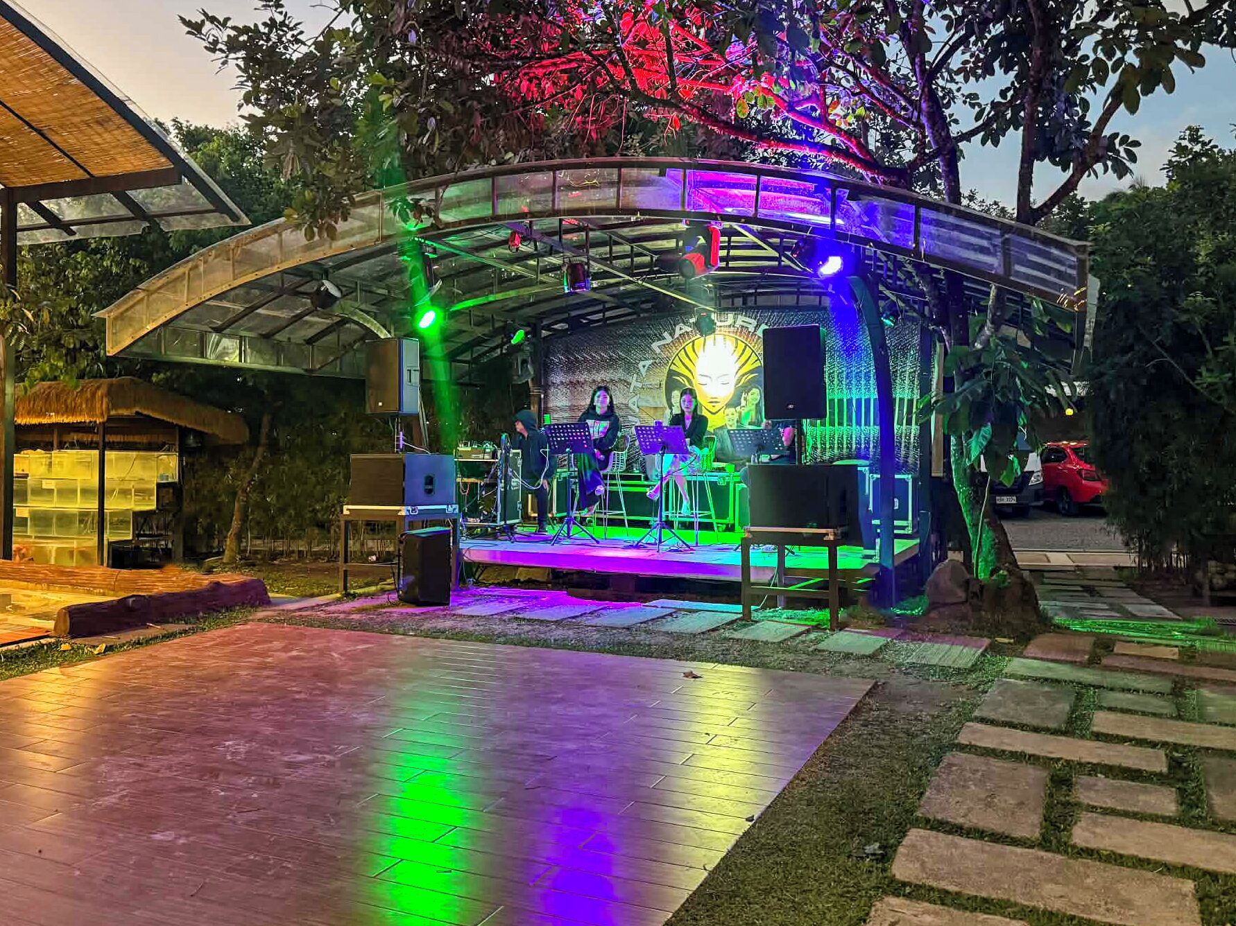 Un escenario al aire libre con equipo musical e intérpretes, rodeado de árboles y una zona cubierta.