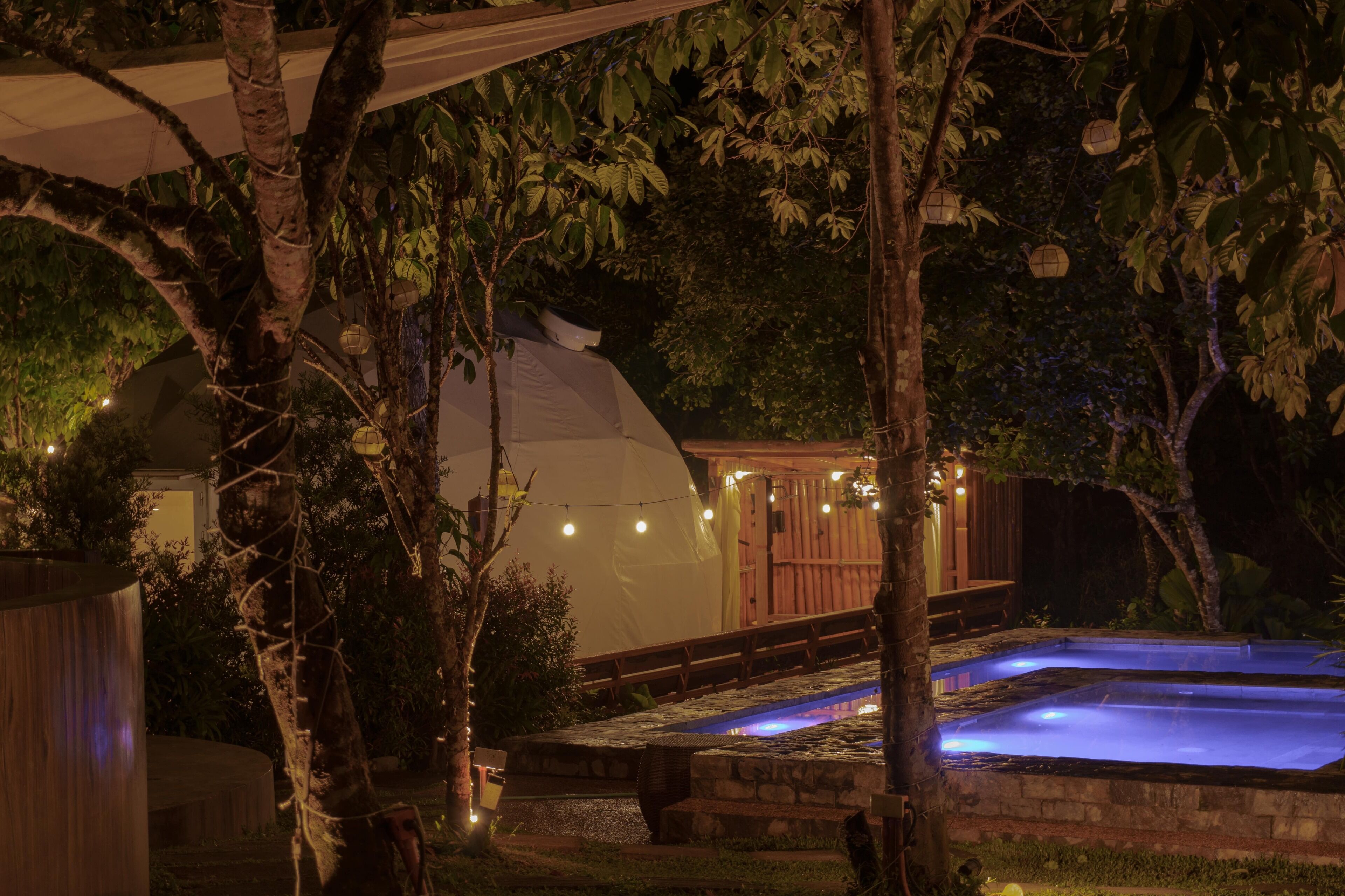 A swimming pool with a wooden deck, surrounded by trees and a covered area with string lights.