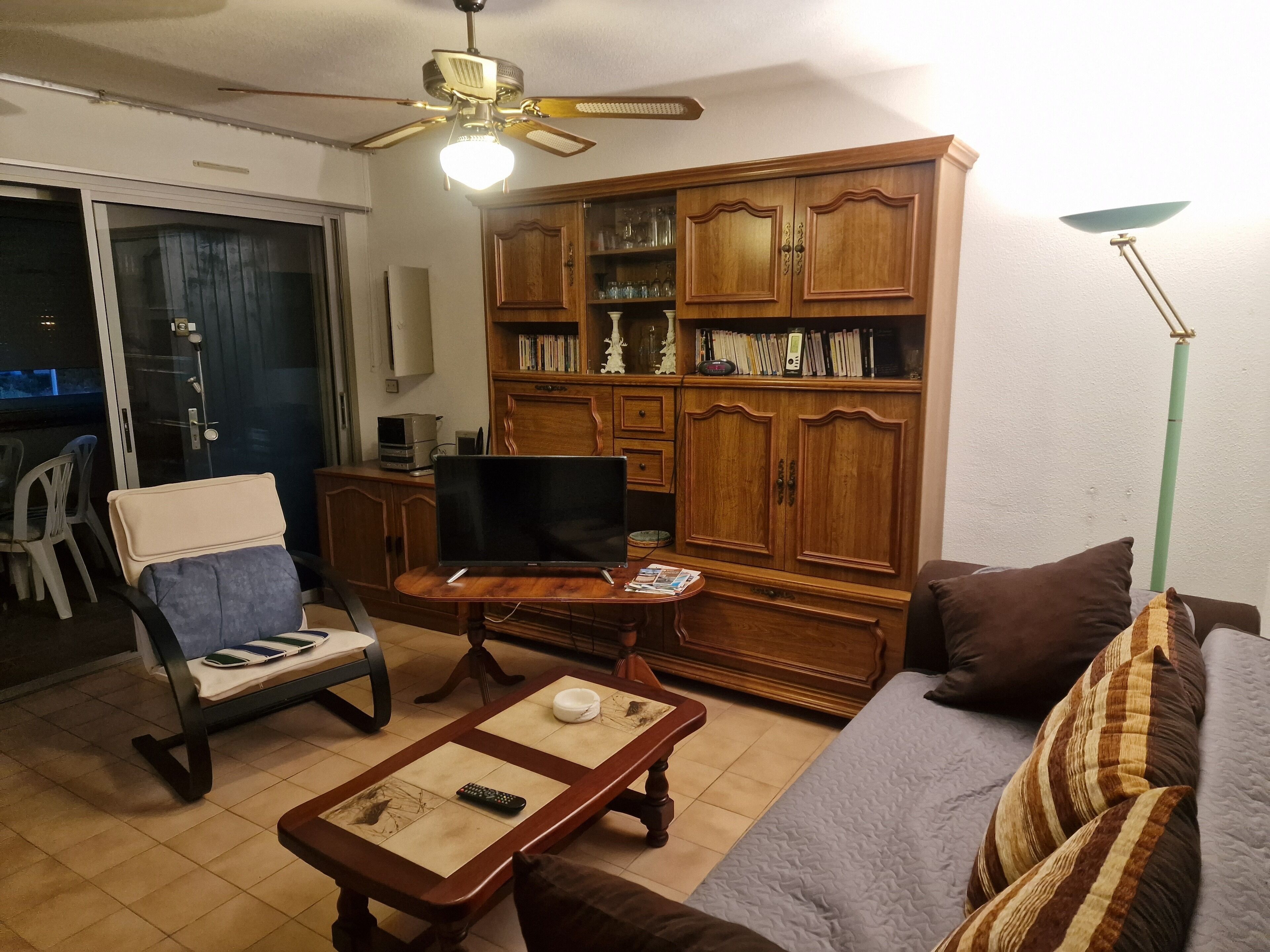 A living room with a sofa, armchair, coffee table, television, wooden cabinet, and a ceiling fan.