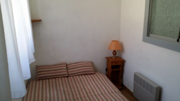 Een kleine, eenvoudige kamer met een bed, een nachtkastje met een lamp en een radiator.