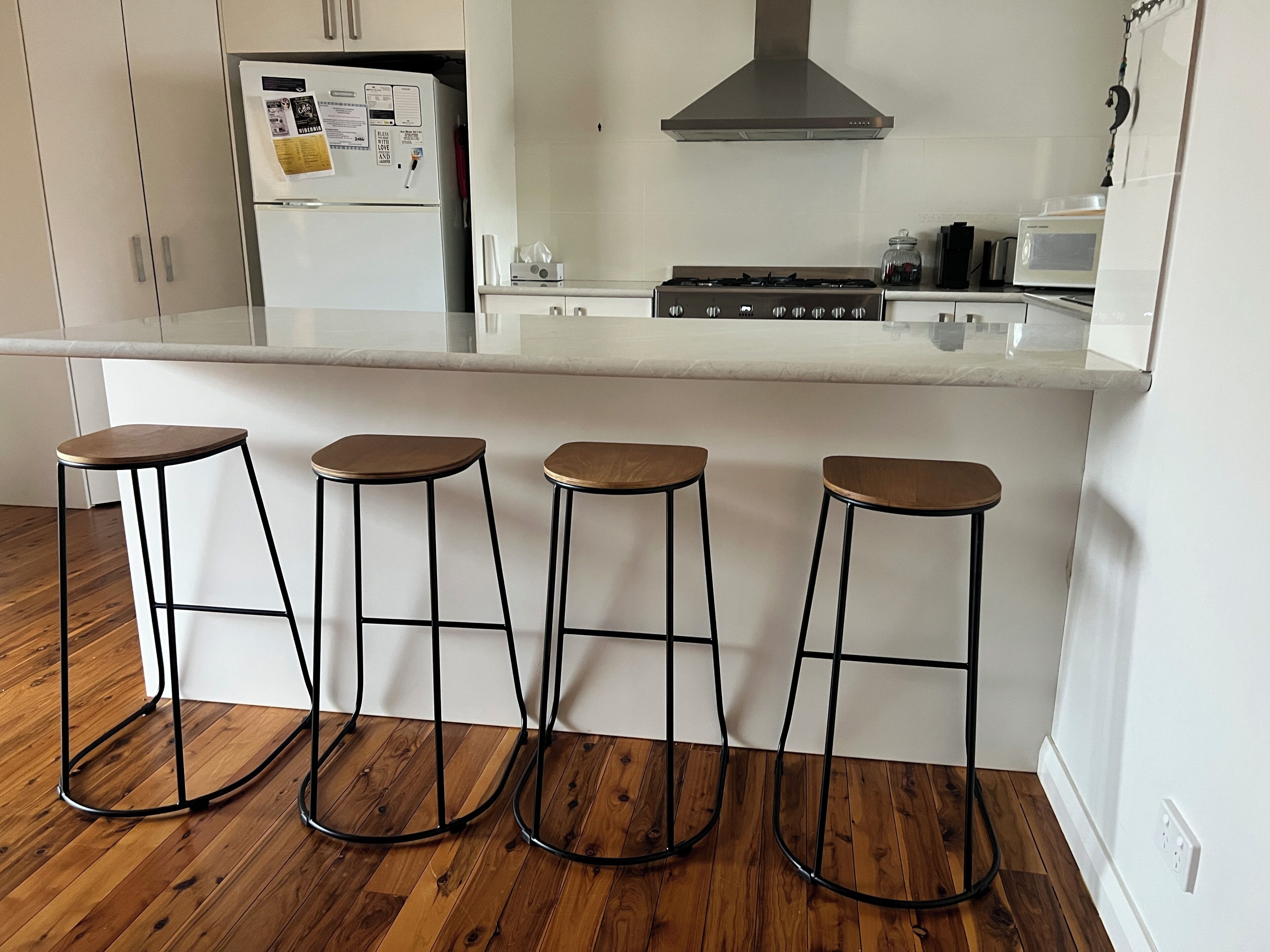 Une cuisine avec un plan de travail blanc, quatre tabourets, une cuisinière et des placards.